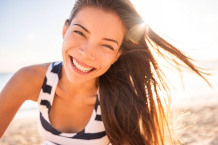 Happy Asian woman smiling in sun flare beach natural beauty model with perfect teeth laughing of joy and fun on summer vacation. Long brown healthy hair on sunlight sunset glow.の写真素材