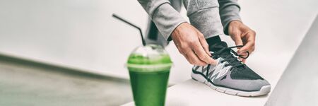 Green juice weight loss smoothie fit man getting ready for cardio run workout tying running shoes laces drinking detox drink panoramic banner. Fitness runner athlete with healthy vegan juice.の写真素材
