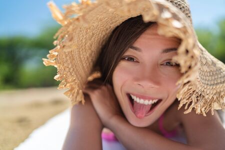 Happy funny face Asian young woman doing goofy facial expression sticking tongue out on summer vacation relax holiday sun tanning on beach having fun.の写真素材