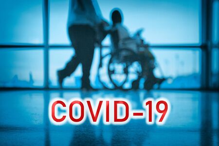 COVID-19 billboard red text on blue medical hospital background with doctor walking with disabled patient in wheelchair.の写真素材