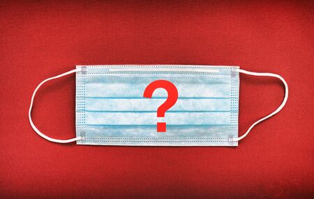 COVID-19 - is it necessary to wear a mask. Should wearing a face mask be compulsory in cities where social distancing is not possible. Question mark on mask red background.の写真素材