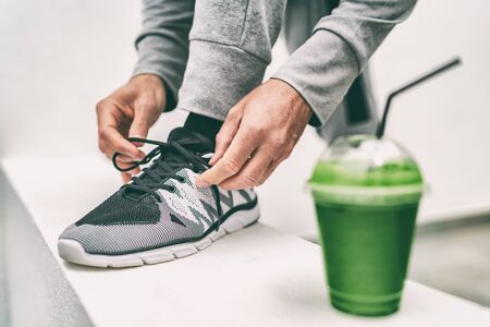 Green juice exercise man getting ready for cardio run workout tying running shoes laces drinking smoothie drink. Fitness runner athlete lacing shoe, with vegetable detox cleanse juice healthy eating.の写真素材