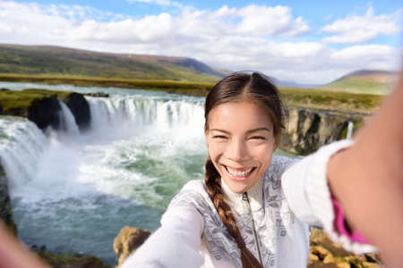 Tourist taking video selfie on travel by Godafoss waterfall on Iceland. Happy young woman tourists enjoying icelandic nature landscape visiting famous tourist destination attraction, Iceland.の写真素材