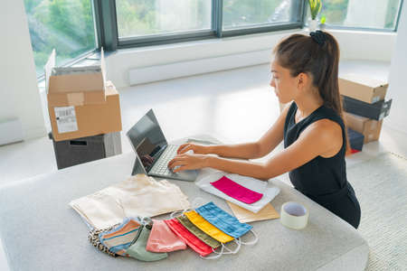 Online small business entrepreneur woman working remote from home on laptop selling cloth mask handmade from fabric on ecommerce app for COVID-19.の写真素材