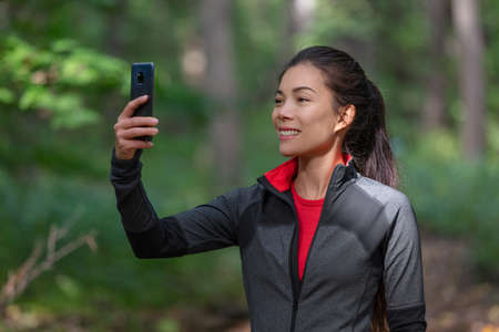 Phone active woman taking picture with mobile cellphone outside on forest nature walk during cardio activity holding smartphone.の写真素材