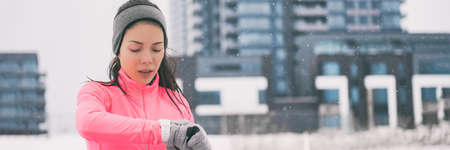 Winter running smartwatch fitness woman in the city. Asian girl in cold snow weather jogging outside...