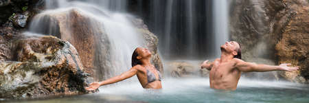 Nature couple relaxing under waterfall with open arms in freedom banner. People enjoying water falling in natural pool in tropical nature. Wellness, health and spa. Wide landscape crop for panorama.の写真素材