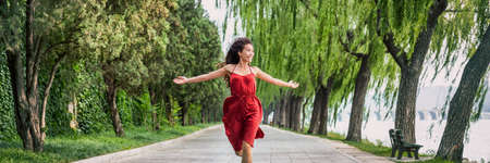 Happy woman running carefree and in freedom enjoying breathing healthy in beautiful summer park banner background. Asian girl enjoying red dress living life. Banner panorama.の写真素材