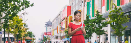 Asian woman walking downtown on Wangfujing shopping street in Beijing, china, using phone app to find directions or text online. Happy Chinese woman texting on mobile sms smartphone.の写真素材