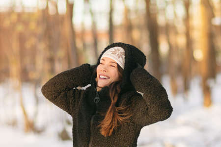 Winter snow fun happy girl walking outside in cold weather protecting ears holding wool hat over ears active outdoor lifestyle. Asian girl wearing black outerwear jacket, gloves breathing fresh air.の写真素材