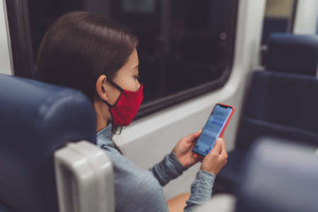 Train commute during corona virus pandemic. Woman passenger using mobile phone wearing face mask sitting in public transport texting online on contact tracing app.の写真素材