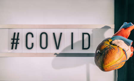 COVID-19 hashtag sign next to medical heart model showing cardiovascular risk of stroke blood clot with coronavirus infection. Lightbox billboard text.の写真素材