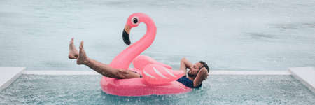 Happy man relaxing in swimming pool flamingo float despite bad rain weather. Travel summer vacation banner.の写真素材