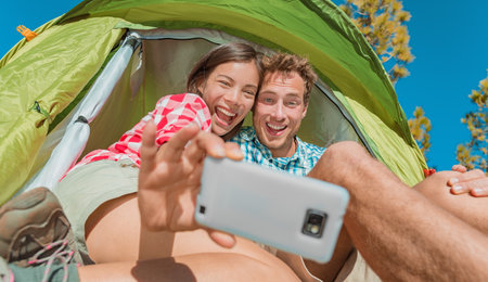 Camping selfie fun couple tourists holding phone taking self portrait photos in tent during summer vacation travel. Asian girl, Caucasian man laughing doing silly facesの写真素材