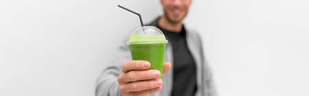 Green smoothie juice drink at cafe banner. Healthy man drinking detox diet protein shake white background panoramic. Hand holding plastic cup of vegetable juice, health food eating lifestyle.の写真素材
