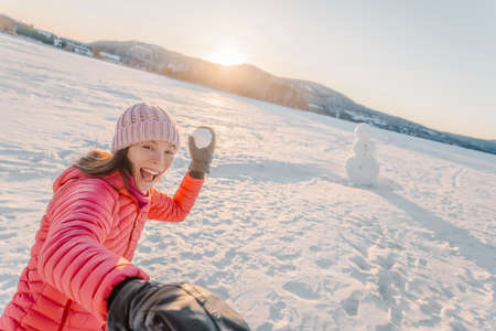 Winter girl throwing snowball at camera smiling happy having fun outside on snowy winter day playing...