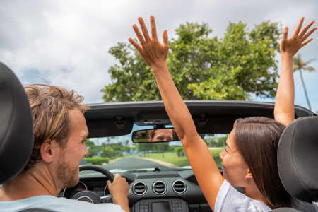 Happy people driving new convertible car on road trip. Asian woman with arms up laughing with boyfriend through open roof having fun driving on summer vacation travelの写真素材