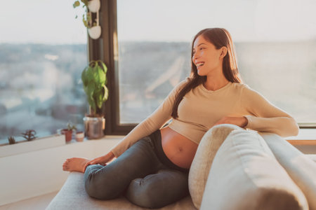 Happy Asian pregnant woman relaxing at home watching tv on sofa in living room. Laughing young girl during pregnancyの写真素材