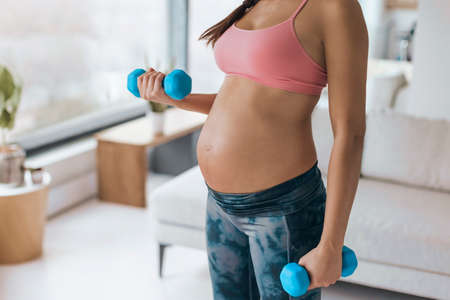 Pregnant woman home workout. fitness strength training exercise with dumbbell weights. Body workout arm curls closeup of belly and armsの写真素材