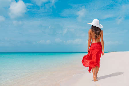 Luxury beach vacation elegant tourist woman walking relaxing in red beachwear and sunhat on white sand Caribbean beach. Lady tourist on holiday vacation resortの写真素材