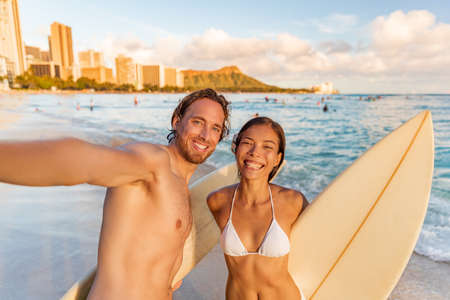 Hawaii beach selfie happy couple surfers smiling to camera taking phone photo at surfing lesson in Waikiki, Honolulu city, Oahu island, Hawaii, USA. Asian bikini woman holding surfboard on vacationの写真素材