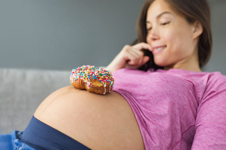 Unhealthy food during pregnancy. Sugar temptation craving Asian pregnant woman trying to resist eating a sweet donut on her baby belly. Funny eating cravings conceptの写真素材