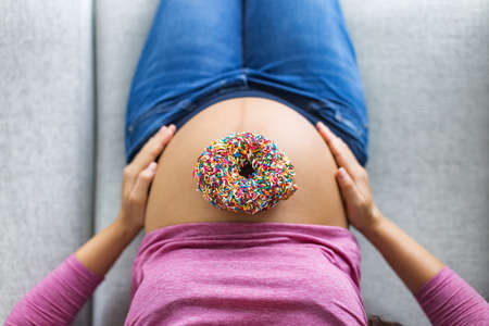 Pregnant woman with donut on belly top view. Cravings of desserts and sweets during pregnancy, Pastry with birthday cake sprinkles on baby bump for gestational diabetesの写真素材