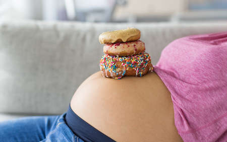 Pregnant woman with donuts on belly. Cravings of desserts and sweets during pregnancy, Girl eating unhealthy pastries on baby bump for gestational diabetesの写真素材