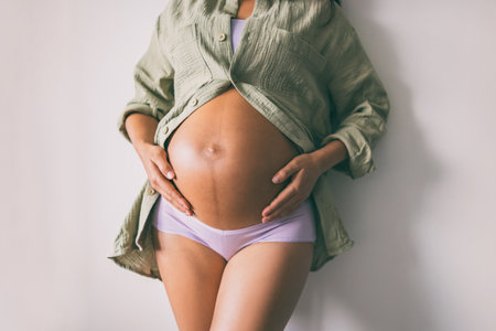 Pregnancy belly closeup. Pregnant woman wearing underwear and casual cotton shirt at home relaxing holding expecting tummy for skincare, health, lifestyleの写真素材