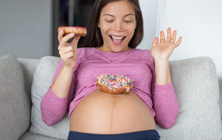 Pregnancy food cravings concept photo. Funny Asian pregnant woman eating donuts at home hungry for dessert cakes and sweet snacks. Gestational diabetes, nutrition, weight gainの写真素材