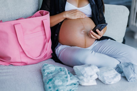 Pregnant woman preparing hospital bag for birth for mom and baby while looking at phone for maternity hospital bag checklist and online shopping. Expecting woman packing diapers, baby clothing etcの写真素材