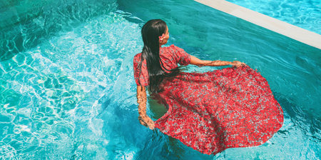Summer dress fashion model woman swimming in pool wearing long floral red maxi dress clothing. Creative artistic fashion photo shoot at luxury resort infinity poolの写真素材
