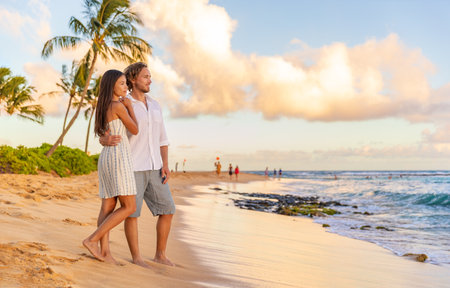 Couple on romantic sunset beach walk relaxing during Hawaii summer travel vacation on Kauai island, USA. Asian woman in white dress, Man in linen shirtの写真素材