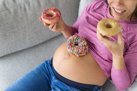 Pregnant woman eating sweet donuts on belly. Cravings of desserts and unhealthy food during pregnancy, Funny top view of cakes on baby bump for gestational diabetes or food cravings during pregnancyの写真素材