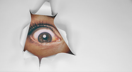 Eye peeking through peep hole watching something scary. Scared woman with look of horror in her eyes looking peeping from a dark room at nightの写真素材