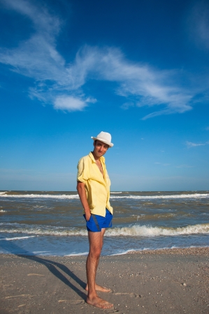 Happy young man in hat, blue shorts and yellow shirt standing on the beach near the sea and smiling. Summer moodの写真素材