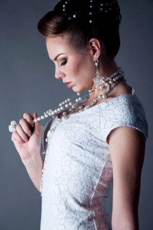 Studio portrait of beautiful young brunette bride all in white wearing white wedding dress, tender makeup, elegant hairdo and a lot of jewelry  like pearl necklace, crystal glass earrings, rings and looking at her handの写真素材