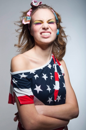 Studio portrait of young patriotic American girl wearing bright makeup, colorful head band, off the shoulder top tank woth USA flag, making apes and grinning at camera with toothy smile. Gray backgroundの写真素材