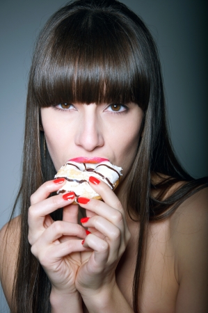 Beauty studio shot of sexy brunette female model with long straight healthy hair, glossy red lips, holding tasty donut with her hands, eating it with great pleasure and temptation, looking at camera. Black backgroundの写真素材