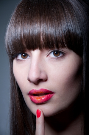 Beauty studio portrait of beautiful young woman model with long straight fringe, natural makeup, bright colorful red glossy lips and nail polish of the same color, holding her finger near lips, looking at camera. Gray background, copy spaceの写真素材