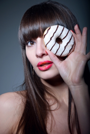 Pretty sexy brunette girl with long straight hair, bare shoulders, glossy red lips, holding tasty donut with hand as a monocle, looking at camera  Dark gray background, copy spaceの写真素材