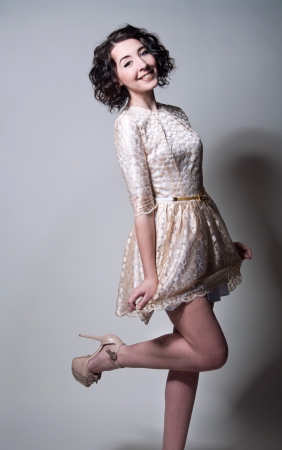 Studio portrait on gray of a beautiful young brunette woman model with curly hair, tender makeup wearing light golden color dress with narrow belt, beige high heels, holding her dress, smiling, looking at cameraの写真素材
