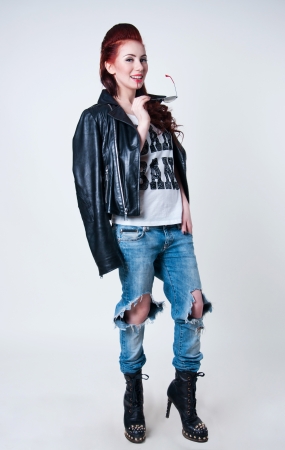 Pretty young rocker woman model with cute red haircut, wearing leather jacket, ripped blue jeans, black boots with high heels and spikes, holding her glasses, looking at camera. Gray background, copy spaceの写真素材
