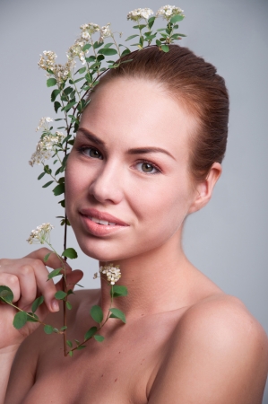 Beauty studio portrait of young beautiful woman model with perfect skin, light natural makeup, holding  blossoming white flowers of spiraea on a branch, looking at camera. Spring mood, gray background, copy spaceの写真素材