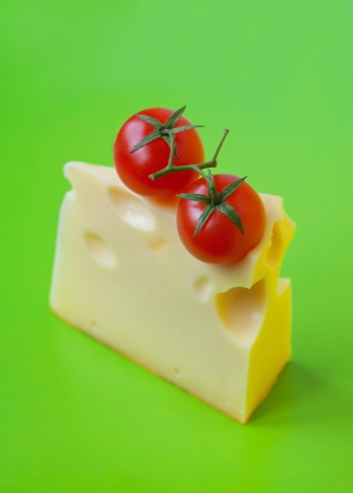 Bright colorful tasty and delicious still life made of two red ripe fresh cherry tomatoes with stems on top of a piece of cheese with big internal holes against bright green background  Copy spaceの写真素材