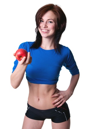 Pretty slim and fit young sporty brawny woman model wearing blue top showing her torso, black shorts, resting after training, holding a ripe red apple in her hand, offering it to a viewer, smiling at camera with toothy smile. Isolated on white, copy spaceの写真素材