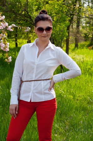Elegant young beautiful woman model in sun glasses, wearing white shirt, red trousers, holding her hand on waist, against fresh green grass and blooming spring trees backgroundの写真素材