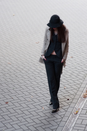 Pretty brunette woman model wearing stylish black pants suit and gray rain coat, black hat with wide fields, high heels, red lips, hands in pockets, going down the street covered with pavement tiles  Copy spaceの写真素材