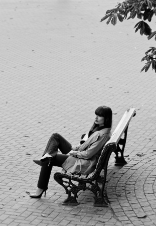 Black and white portrait of pretty brunette woman model wearing stylish black pants suit and gray rain coat, high heels, sitting on a bench with legs crossed high  In autumn on the street covered with pavement tiles  Copy spaceの写真素材