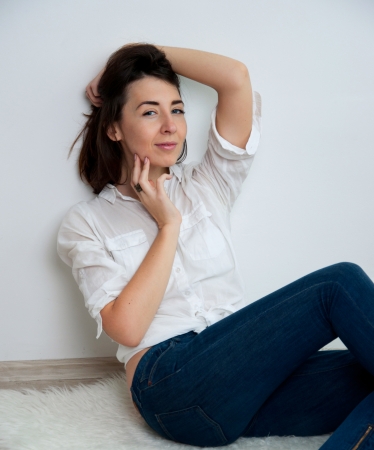 Pretty brunette woman wearing blue jeans, white shirt, sitting on the floor in a minimalistic room against white wall, looking at camera, smiling. Copy spaceの写真素材